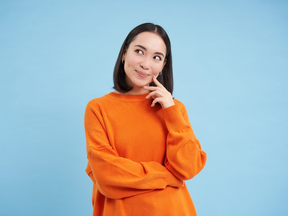 Smiling thinking asian woman, looking up with thoughtful, pleased face expression, has interesting idea, plans something, standing over blue background